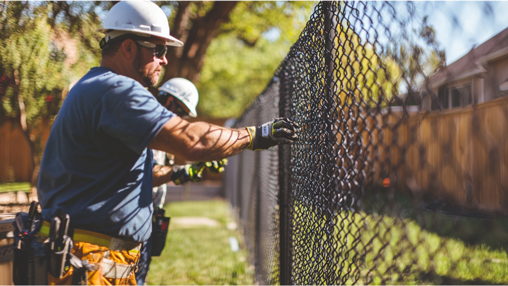 Construction workers installing a black chain-link fence in a residential neighborhood on a sunny day.