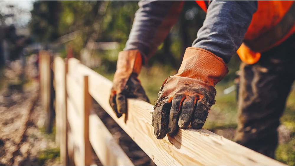 Close-up of a construction worker wearing orange gloves installing a wooden fence in an outdoor setting.